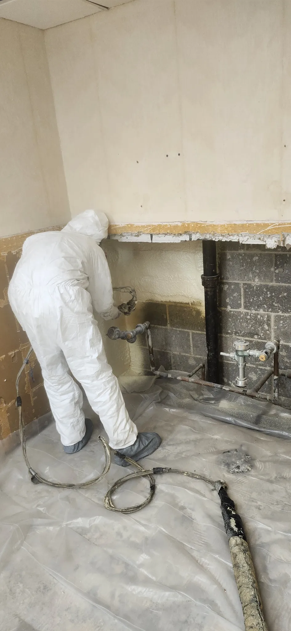 Worker spraying foam behind plumbing pipes in commercial renovation